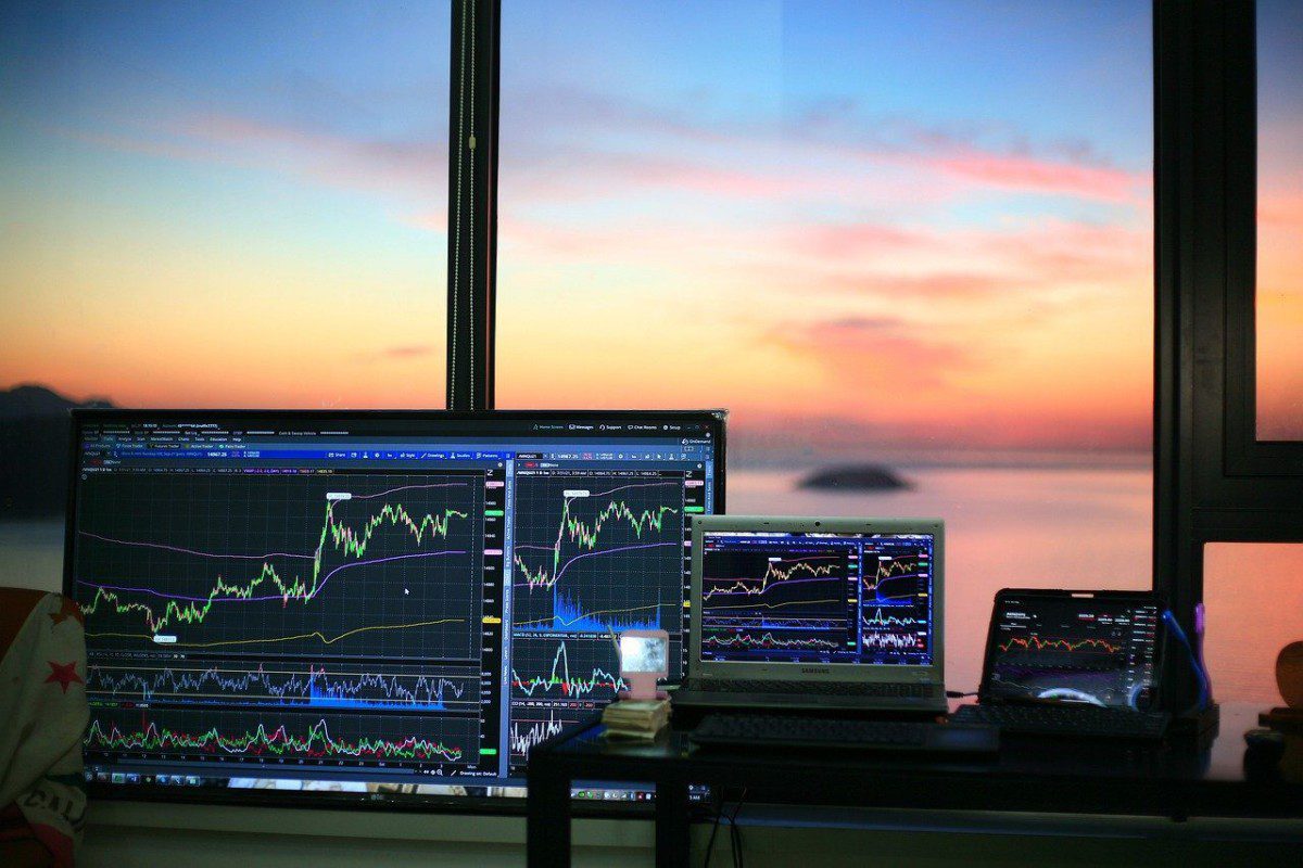 A imagem mostra um monitor, um notebook e um tablet, que exibem gráficos da variação do preço de ações. As telas estão em frente a uma janela e pode-se ver o pôr do sol ao fundo.