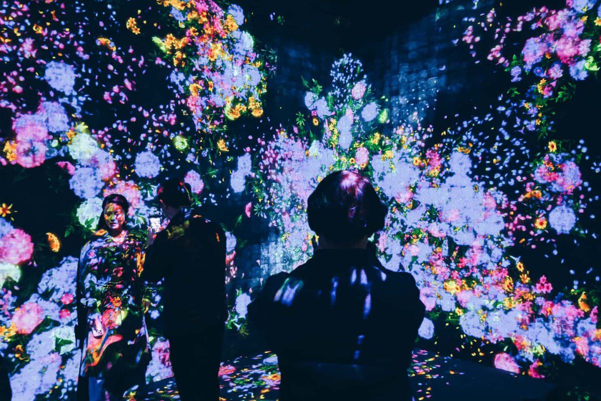 A imagem mostra pessoas em uma exposição de arte digital. O ambiente está escuro. A obra é exibida em telões e é composta por flores de diversos tipos e cores.
