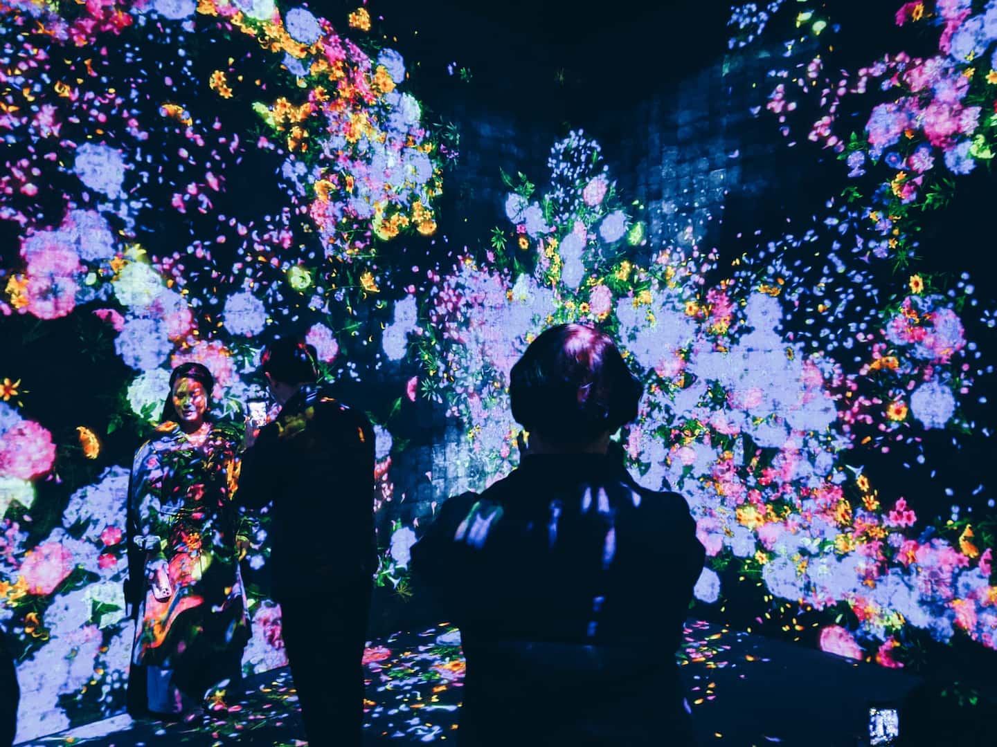 A imagem mostra pessoas em uma exposição de arte digital. O ambiente está escuro. A obra é exibida em telões e é composta por flores de diversos tipos e cores.