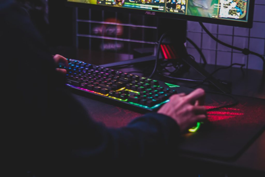 Pessoa jogando em um computador gamer, com teclado e mouse com luzes coloridas. É possível ver apenas o braço e as mãos da pessoa, que usa um moletom preto.