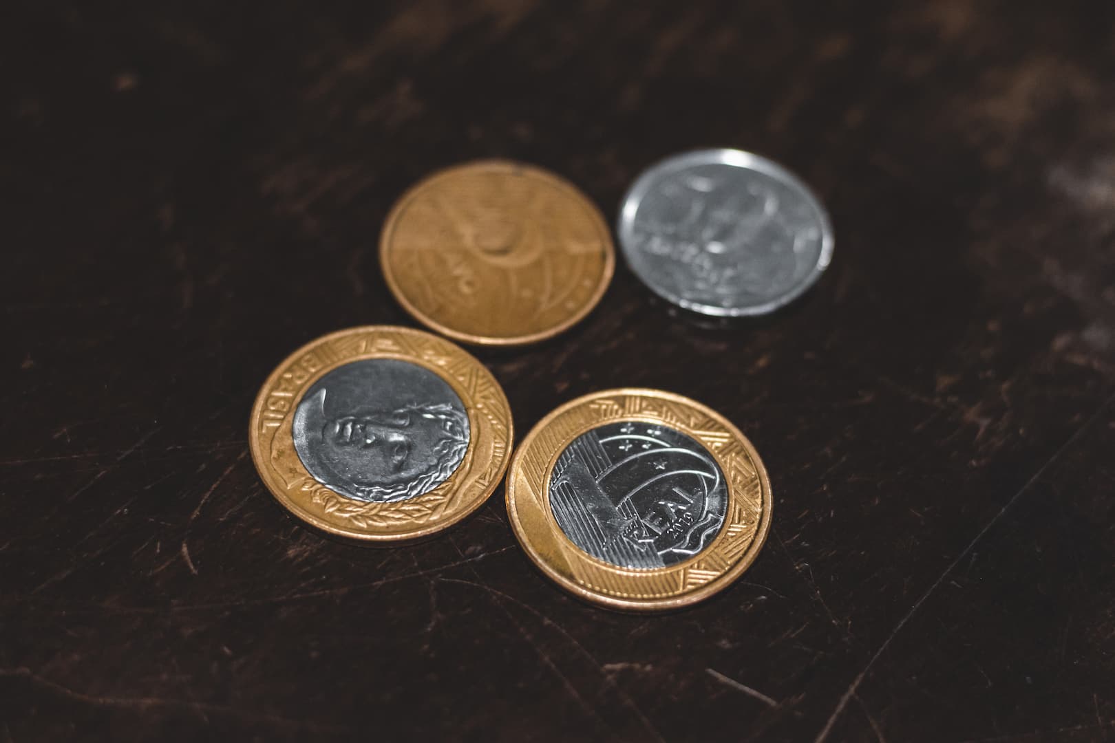 A imagem mostra quatro moedas em cima de uma superfície de madeira escura. Há duas moedas de um real, uma de cinquenta centavos e uma de 25 centavos.