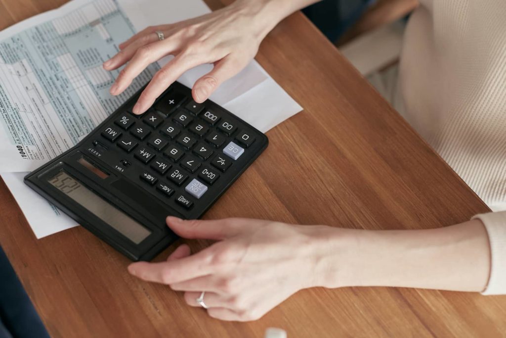 Pessoa branca calculando o valor do ICMS com uma calculadora preta em cima de uma mesa de madeira.