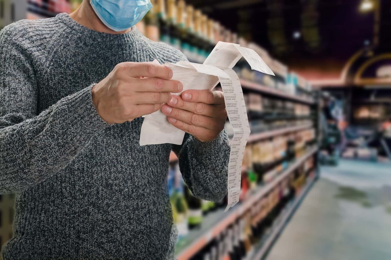 Homem branco, usando uma blusa de frio cinza e uma máscara azul, está em um supermercado lendo a nota fiscal de sua compra.