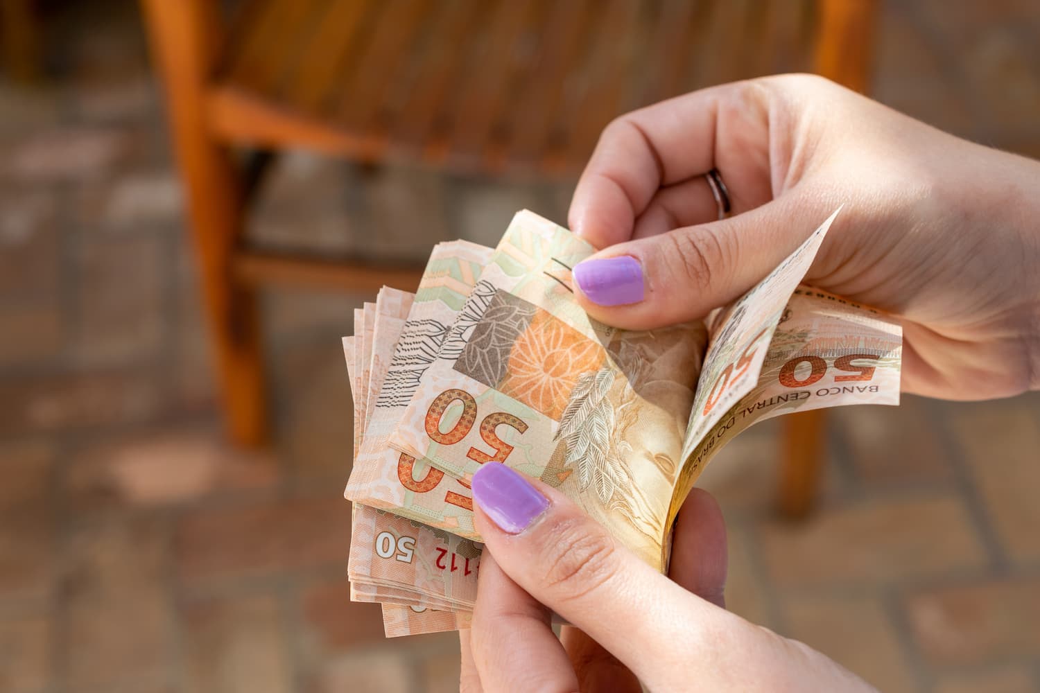 A imagem mostra as mãos de uma pessoa branca, com unhas pintadas de lilás, que está contando notas de 50 reais como exemplo do dissídio salarial. (Foto:Shutterstock / Leonidas Santana)