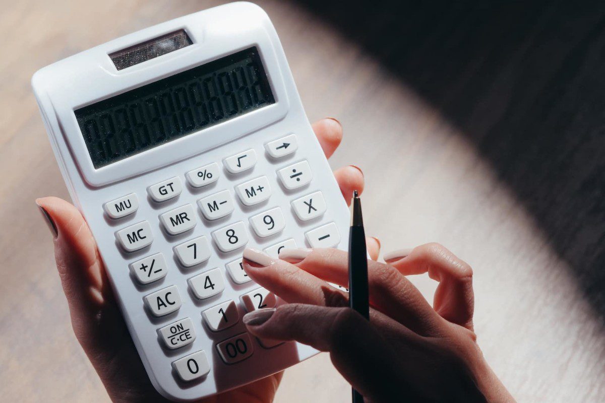 A imagem mostra um par de mãos brancas, com as unhas pintadas, fazendo contas em uma calculadora branca. (Foto: Pexels / Kindel Media)