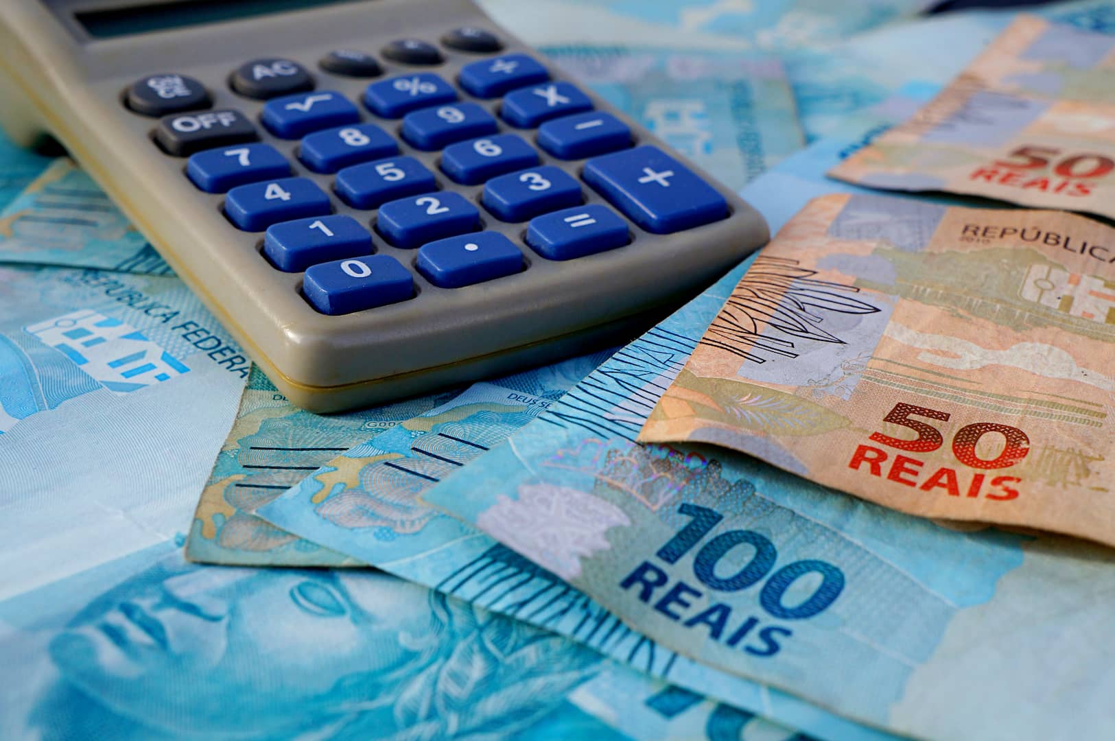 A imagem mostra uma calculadora cinza com botões azuis em cima de notas de R$50 e R$100. (Foto: Shutterstock / Brenda Rocha)