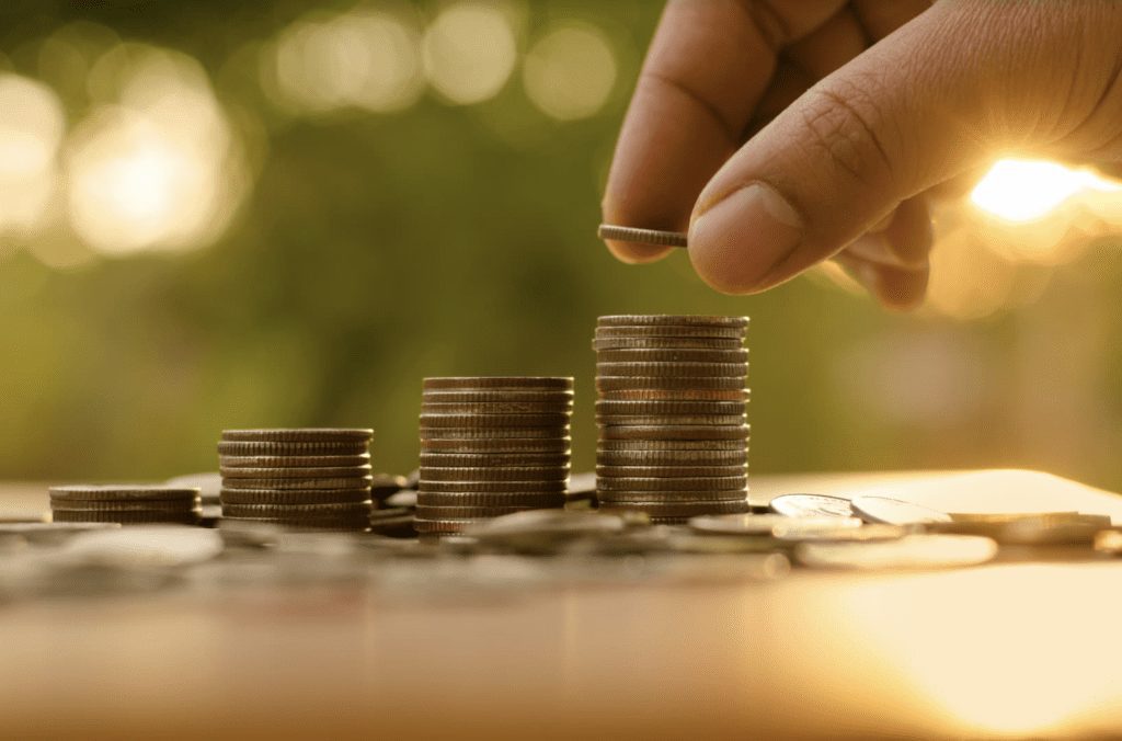 A imagem mostra a mão de uma pessoa branca adicionando uma moeda em cima de uma pilha de moedas. (Foto: Shutterstock / Singkham)