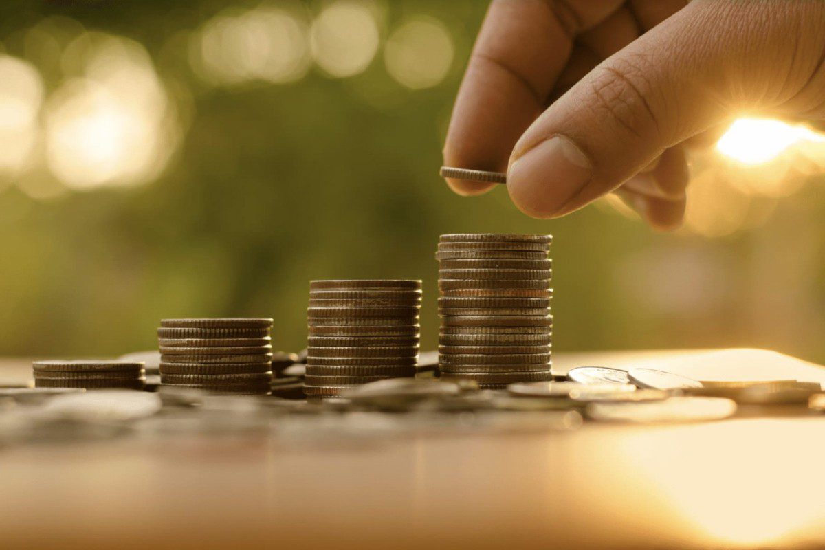 A imagem mostra a mão de uma pessoa branca adicionando uma moeda em cima de uma pilha de moedas. (Foto: Shutterstock / Singkham)