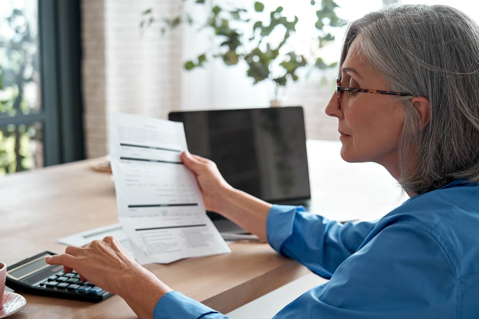 A imagem mostra uma mulher idosa sentada em frente a uma mesa de madeira. Ela segura um documento com dados em uma mão, enquanto faz contas em uma calculadora. (Foto: Shutterstock / Ground Picture)