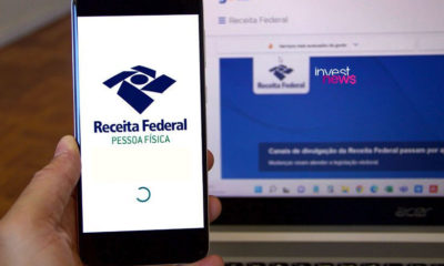 uma mão segurando um smartphone com a tela mostrando o aplicativo da Receita Federal. Ao lado, um notebook mostrando o site da Receita Federal