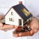 Foto de uma miniatura de uma casa segurada na palma de duas mãos, ao lado de uma chave, ilustrando o "Minha Casa, Minha Vida"