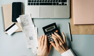 Mão de mulher calculando, usando calculadora, em mesa com notebook, anotações e notas fiscais