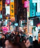 Pessoas andando entre prédios em uma rua iluminada à noite; Seoul, Coreia do Sul