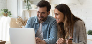 casal de homem e mulher sorrindo em frente a um notebook com tela aberta. na mesa há um caderno e uma calculadora