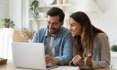 casal de homem e mulher sorrindo em frente a um notebook com tela aberta. na mesa há um caderno e uma calculadora