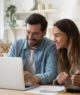 casal de homem e mulher sorrindo em frente a um notebook com tela aberta. na mesa há um caderno e uma calculadora