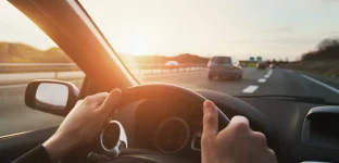 Close do painel de um carro. Em primeiro plano, vê-se o close de duas mãos ao volante e ao fundo uma autoestrada com carros circulando