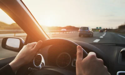 Close do painel de um carro. Em primeiro plano, vê-se o close de duas mãos ao volante e ao fundo uma autoestrada com carros circulando