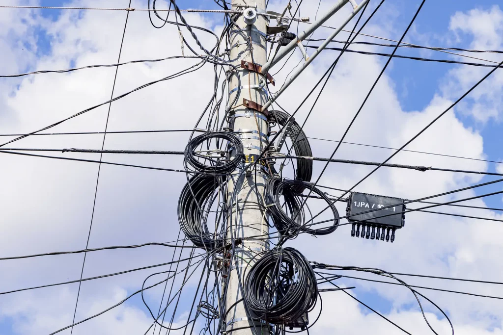 Emaranhado de cabos de energia elétrica presos no alto de um poste de concreto com um céu azul com nuvens ao fundo