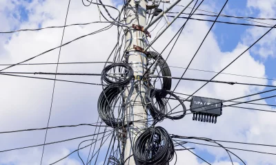 Emaranhado de cabos de energia elétrica presos no alto de um poste de concreto com um céu azul com nuvens ao fundo