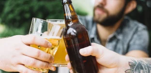 Detalhe das mãos de pessoas brindando com copos e garrafa de cerveja, com um homem desfocado ao fundo