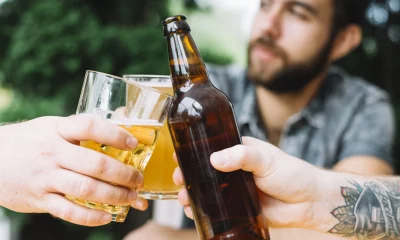 Detalhe das mãos de pessoas brindando com copos e garrafa de cerveja, com um homem desfocado ao fundo