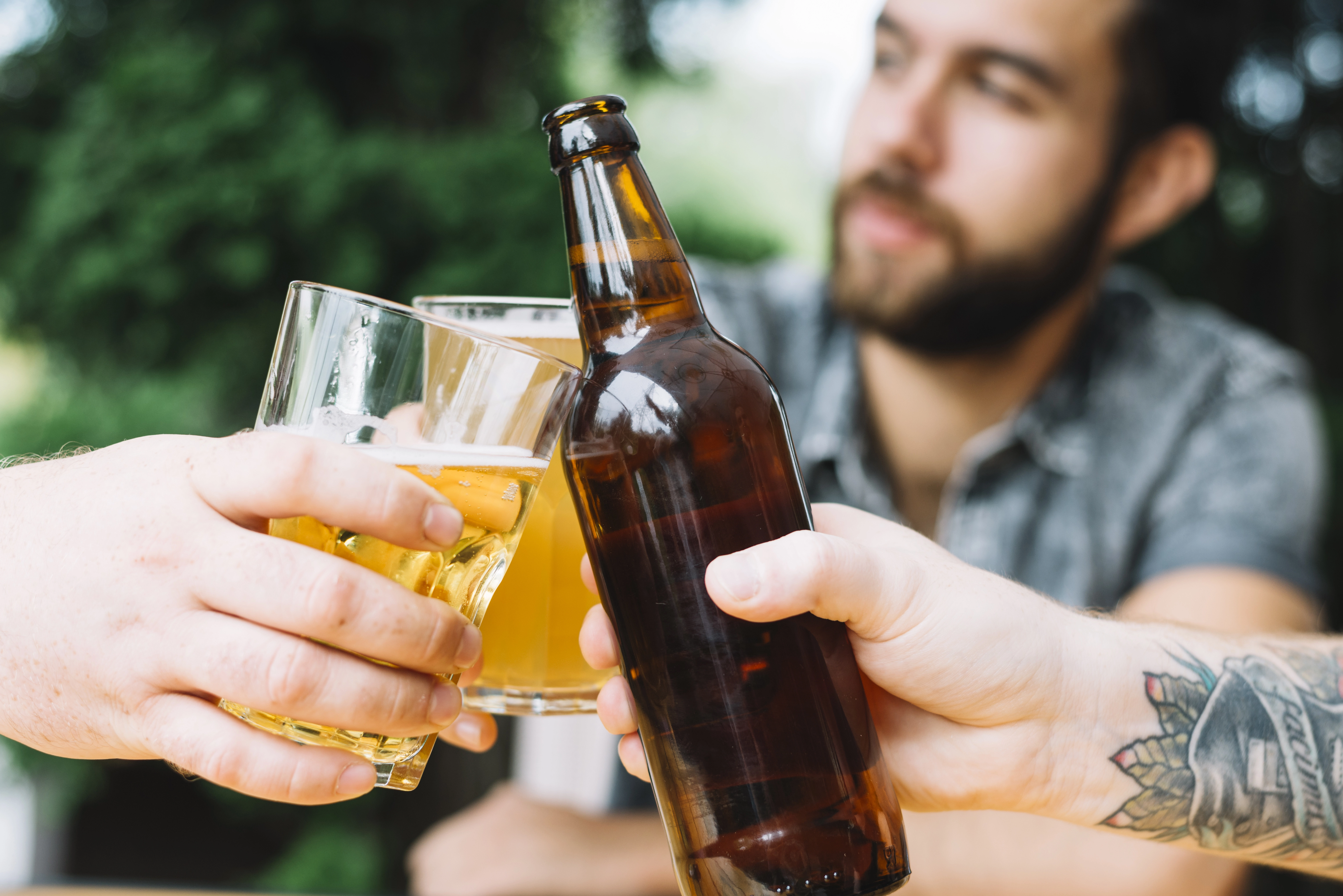 Detalhe das mãos de pessoas brindando com copos e garrafa de cerveja, com um homem desfocado ao fundo