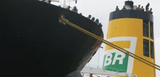 Recorte de um navio petroleiro com trabalhadores na borda do convés, observando o porto. Em segundo plano, uma chaminé amarela com o logo da BR distribuidora.