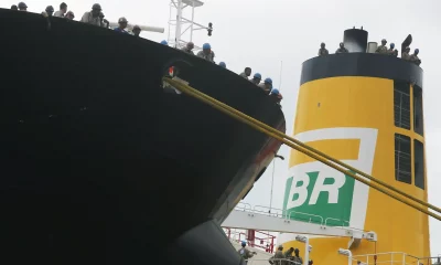 Recorte de um navio petroleiro com trabalhadores na borda do convés, observando o porto. Em segundo plano, uma chaminé amarela com o logo da BR distribuidora.