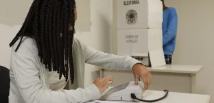 Pessoa vota em urna eletrônica na cabine de votação, enquanto mesária aguarda para entregar o comprovante de votação