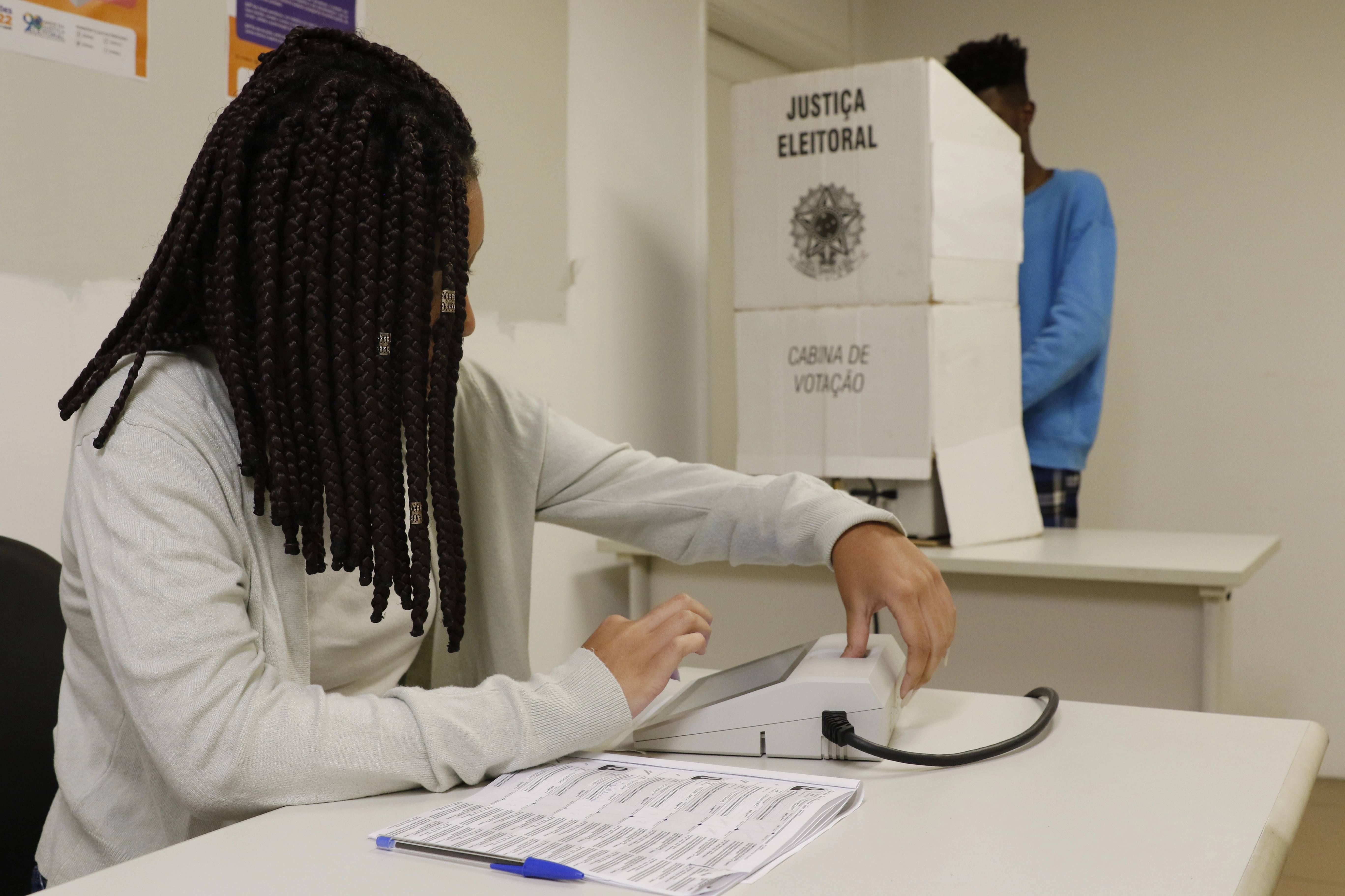 Pessoa vota em urna eletrônica na cabine de votação, enquanto mesária aguarda para entregar o comprovante de votação