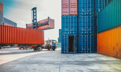 Imagem de um movimentado pátio de contêineres, com diversas pilhas coloridas, um caminhão e um reach stacker em operação sob um céu parcialmente ensolarado.