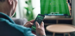 Pessoa de costas e de perfil segurando um celular com aplicativo de apostas esportivas, enquanto uma TV ao fundo exibe uma partida de futebol ao vivo, em um ambiente interno aconchegante com mobiliário moderno.