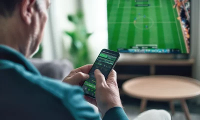 Pessoa de costas e de perfil segurando um celular com aplicativo de apostas esportivas, enquanto uma TV ao fundo exibe uma partida de futebol ao vivo, em um ambiente interno aconchegante com mobiliário moderno.