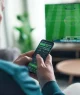 Pessoa de costas e de perfil segurando um celular com aplicativo de apostas esportivas, enquanto uma TV ao fundo exibe uma partida de futebol ao vivo, em um ambiente interno aconchegante com mobiliário moderno.