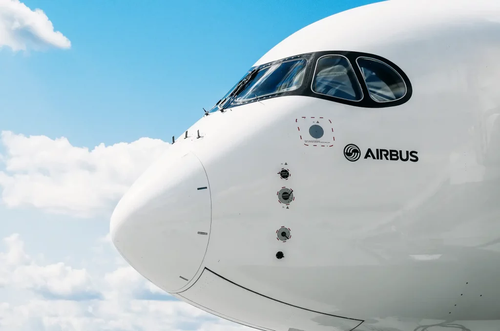 Parte frontal de uma aeronave da Airbus contra um céu azul com algumas nuvens esparsas, mostrando o nariz do avião, a cabine de comando com janelas curvas e o logotipo da Airbus na fuselagem branca.