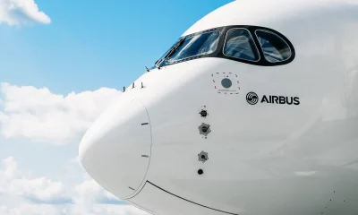 Parte frontal de uma aeronave da Airbus contra um céu azul com algumas nuvens esparsas, mostrando o nariz do avião, a cabine de comando com janelas curvas e o logotipo da Airbus na fuselagem branca.