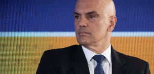 Retrato do ministro do Supremo Tribunal Federal (STF) Alexandre de Moraes, um homem branco e careca, de terno preto, camisa branca e gravata azul escura com padrões discretos está em frente a uma tela digital azul e laranja.