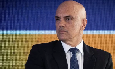 Retrato do ministro do Supremo Tribunal Federal (STF) Alexandre de Moraes, um homem branco e careca, de terno preto, camisa branca e gravata azul escura com padrões discretos está em frente a uma tela digital azul e laranja.