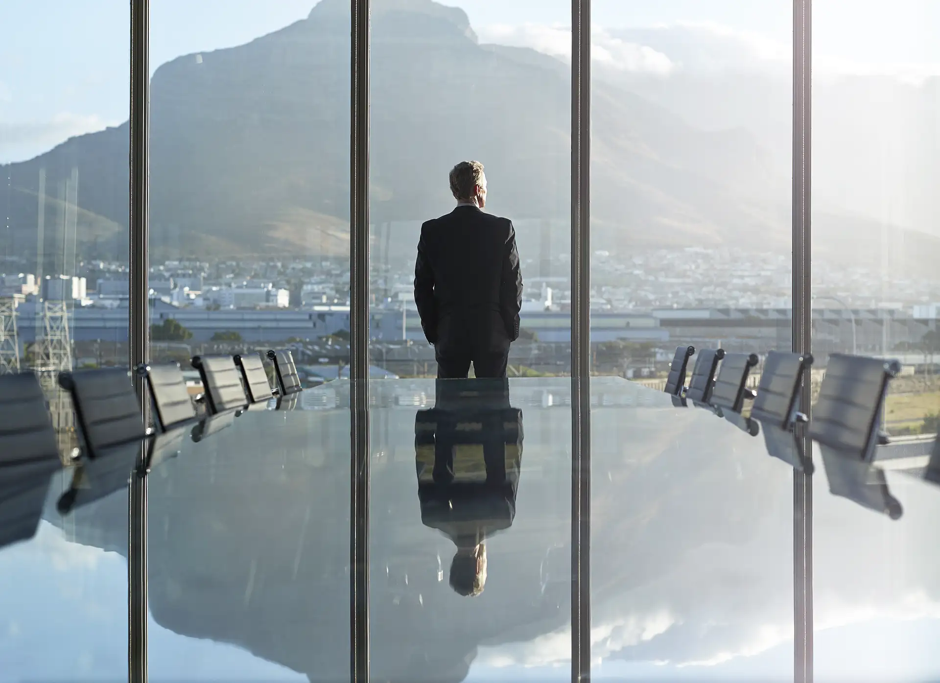 Um homem de costas em um terno preto, olhando pela janela de uma moderna sala de reuniões, com vista para uma cidade e montanhas ao fundo, em um momento contemplativo.