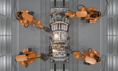 Linha de produção automotiva com braços robóticos laranja montando e soldando a estrutura de um carro.