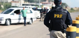 Na imagem, um policial da PRF de costas, com uniforme preto e inscrições amarelas, observa uma operação de fiscalização em uma estrada com barreiras, veículos parados e outros policiais. Ao fundo, construções e uma área verde sugerem tráfego na região.