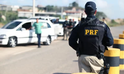 Na imagem, um policial da PRF de costas, com uniforme preto e inscrições amarelas, observa uma operação de fiscalização em uma estrada com barreiras, veículos parados e outros policiais. Ao fundo, construções e uma área verde sugerem tráfego na região.