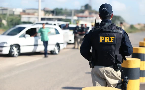 Na imagem, um policial da PRF de costas, com uniforme preto e inscrições amarelas, observa uma operação de fiscalização em uma estrada com barreiras, veículos parados e outros policiais. Ao fundo, construções e uma área verde sugerem tráfego na região.