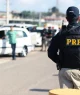 Na imagem, um policial da PRF de costas, com uniforme preto e inscrições amarelas, observa uma operação de fiscalização em uma estrada com barreiras, veículos parados e outros policiais. Ao fundo, construções e uma área verde sugerem tráfego na região.