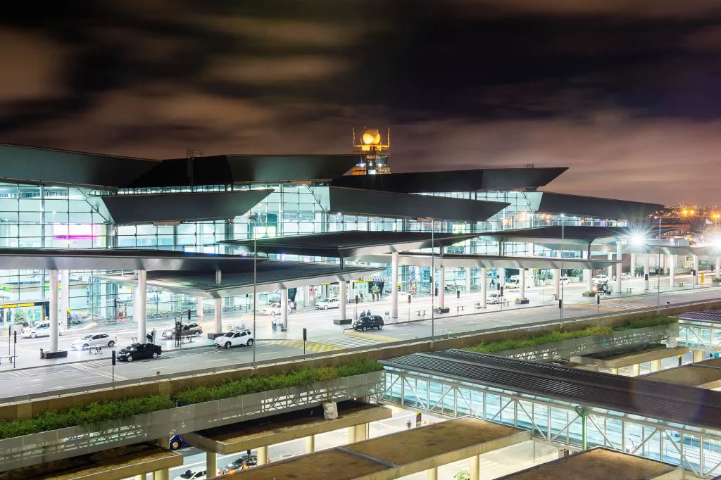 Entrada do Aeroporto de Guarulhos, com estruturas de vidro, torre de controle iluminada, telhado dinâmico, via movimentada e iluminação intensa.