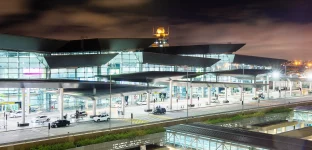 Entrada do Aeroporto de Guarulhos, com estruturas de vidro, torre de controle iluminada, telhado dinâmico, via movimentada e iluminação intensa.