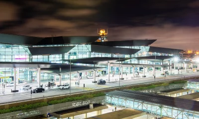 Entrada do Aeroporto de Guarulhos, com estruturas de vidro, torre de controle iluminada, telhado dinâmico, via movimentada e iluminação intensa.