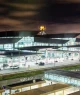 Entrada do Aeroporto de Guarulhos, com estruturas de vidro, torre de controle iluminada, telhado dinâmico, via movimentada e iluminação intensa.