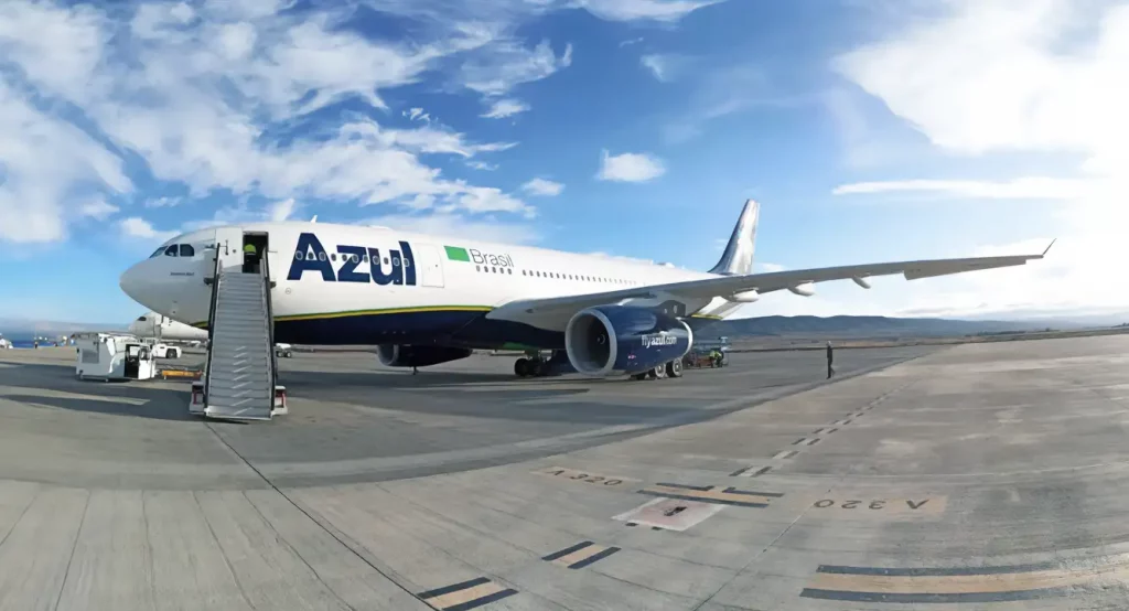 A imagem mostra um avião da companhia aérea Azul estacionado na pista de um aeroporto em um dia claro, com céu azul e algumas nuvens.